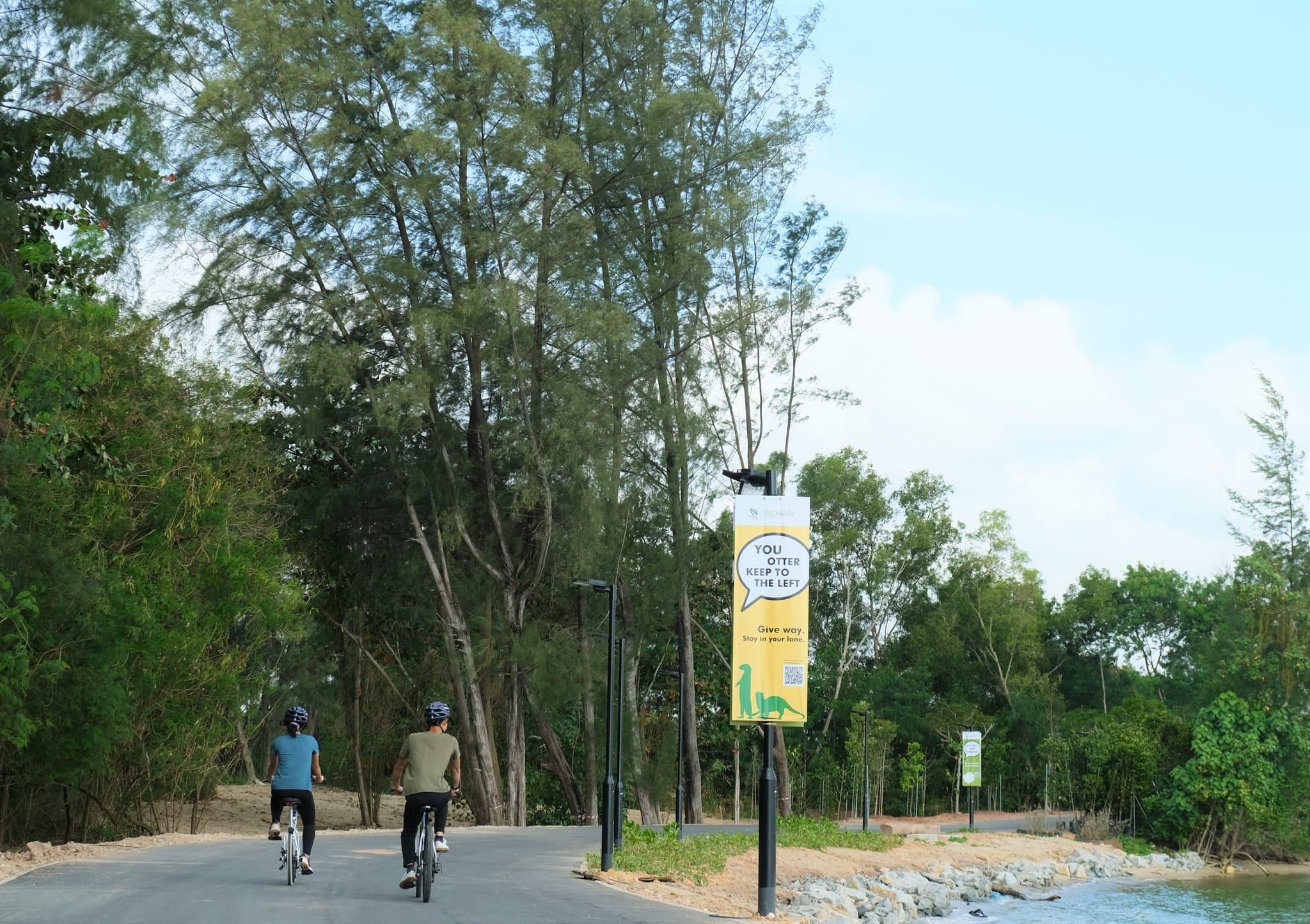 Two cyclists ride on a paved path beside water and trees; a sign reads You otter keep to the left.